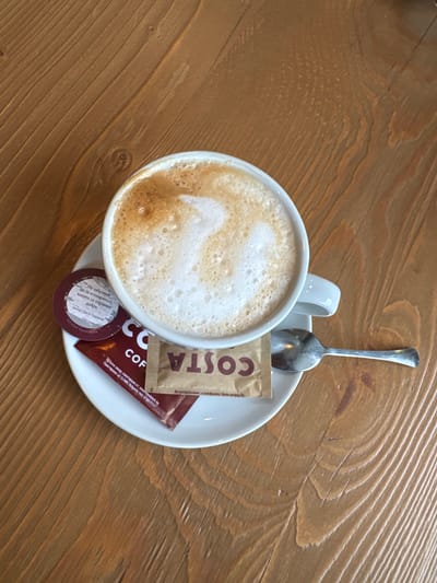 The image shows a cup of coffee on a white saucer, along with a silver spoon and several sugar packets. The cup appears to be a latte or cappuccino, with a foamy surface. One sugar packet is branded with "COSTA COF". A small, round packet contains Bulgarian text. The scene is set on a wooden table with a light brown, textured surface. No people are visible. There's nothing that explicitly indicates the location is Pirdop, Bulgaria, apart from the Bulgarian text on the sugar packet.