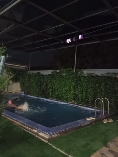 A man is swimming in a pool at night. The pool is surrounded by green grass and a fence covered in ivy. A metal ladder leads into the pool. There are also two pairs of flip-flops near the pool. A covered structure with a metal roof is overhead. Clothes pins are hanging on a wire above the pool. The scene appears to be in a backyard. The location is identified as Sasbaga, Uzbekistan.