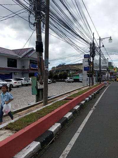 The scene depicts a street view in the city of Taohom, Laos, under an overcast sky. In the immediate foreground, a young girl, wearing a light blue short-sleeved shirt and dark trousers, walks from left to right on a grey paver sidewalk. She holds a white plastic bag, containing what appears to be pink items, in her left hand.
To her right, a concrete curb system separates the sidewalk from an asphalt road. This system consists of a red-painted top section containing green grass, with the lower curb painted in alternating black and white stripes. The asphalt road to the right features a faded white solid line.
Prominently positioned in the mid-ground is a utility pole, partially wrapped in black material, with a green utility box attached. A dense network of numerous black overhead cables extends from this pole and other utility poles, crisscrossing the sky. Further down the street on the right, another utility pole supports a street lamp with a glass shade.
In the background on the left, a multi-story building with a red tiled roof is visible. A blue banner displaying "BRI" and "ATM" is partially visible on its facade. A paved parking lot in front of this building contains several parked white sedans and SUVs, with a motorcycle visible further back. A freestanding blue sign with "BRI" and "ATM" is also present closer to the street on the right. Additional buildings with various roof types are situated in the deeper background. In the far distance on the right, a yellow vehicle is partially visible on the road.