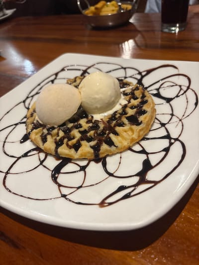 A close-up, high-angle photograph depicts a dessert served on a white rectangular plate with rounded corners, resting on a polished dark wood table within an establishment in Port Alfred, South Africa. The plate features a round, gridded waffle, extensively drizzled with dark chocolate syrup, and topped with two distinct scoops of ice cream: one light beige and one cream-colored. Additional chocolate syrup forms an abstract, swirled pattern across the white plate surface. The blurred background shows a silver bowl containing yellow-orange food items, a clear glass holding a dark beverage, and the upper portion of an individual's light-colored garment.