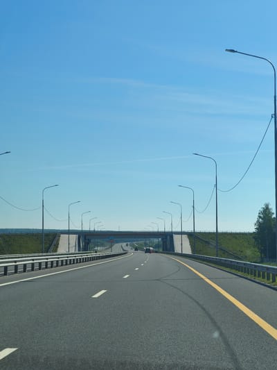 The image shows a highway in Pilekshevo, Russia, on a clear, sunny day. The road is asphalt with clear lane markings, including a solid yellow line on the right and dashed white lines separating lanes. There are guardrails on both sides of the road, and a series of streetlights line the right side of the highway. In the distance, an overpass crosses the highway, with a sign indicating "475". Green grass covers the embankments alongside the road, and a few trees are visible on the right side. Two cars are visible on the highway in the distance. The sky is a bright, clear blue with some faint wispy clouds.