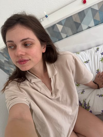 A woman with brown hair and fair skin is lying on a bed with a white pillow patterned with lavender flowers. She is wearing a light beige shirt and a dark bracelet on her left wrist. The bed is near a white wall with a geometric blue and gray design. The location is Kőröshegy, Hungary, as indicated in the source data.
