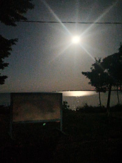 A nighttime scene captures the full moon illuminating a body of water. Silhouetted trees frame the right side of the image, with a dark, shadowy foreground of grass. In the middle ground, a large, rectangular reflective surface stands upright, its surface partially obscured by darkness. The water in the background shows a shimmering reflection of the moonlight and distant lights, suggesting the presence of a shoreline or structures across the water. A power line stretches horizontally across the sky in the upper portion of the image, with the moon's bright light creating visible rays. The location is identified as Durankulak, Bulgaria.