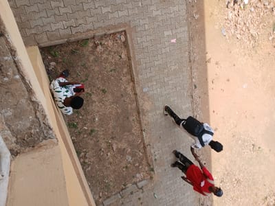 An overhead view captures an outdoor scene in Ugi Majowel, Nigeria. The ground surface consists of a light-colored paved pathway on the left, transitioning to a strip of dry earth with sparse vegetation, and then a larger area of bare, light brown soil on the right. A light-colored building wall with some textured damage occupies the extreme left edge of the frame.
Three individuals are present. One person, with dark hair, is seated on the dry earth patch, leaning against the building wall. This individual wears a white short-sleeved top with an irregular green pattern and dark trousers. They are holding a red rectangular object, possibly a book or tablet, and are looking down at it.
Two other individuals stand on the paved pathway, oriented generally towards the lower-right quadrant of the frame. The first, closer to the center, wears a white t-shirt, a black backpack, and dark trousers. The second, positioned slightly further right and forward, wears a red short-sleeved t-shirt and dark trousers. A distinct linear shadow extends vertically across the right side of the paved area and adjacent dirt.
