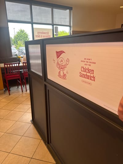 The image shows an indoor scene, possibly within a Chick-fil-A restaurant location in Stone Mountain, Georgia, United States. The foreground features a partition with a prominent advertisement for the Chick-fil-A Chicken Sandwich. The advertisement text reads "We didn't invent the chicken, just the Chicken Sandwich," and "First Made in Atlanta," along with a mascot image. In the background, a window covered in blinds is visible, offering a glimpse of the outside environment, which includes trees and parked cars. Red chairs and tables can be seen through the partition, indicating a seating area. The floor appears to be tiled.