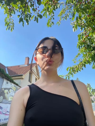 This is a first-person perspective, low-angle photograph taken outdoors on a sunny day. The main subject is a young woman with dark hair wearing sunglasses and a black top. She is positioned in the foreground, with her face angled towards the camera, her lips pursed.

The background reveals a clear blue sky and the branches of a tree with green leaves and small red berries. Below the tree, a building with a tiled roof and brick chimney is visible, suggesting an urban or semi-urban setting. A street sign with some visible text is also present.

The scene appears to be from Dobřany, Czechia. The time of day is likely midday or early afternoon, given the bright sunlight casting shadows and illuminating the scene. The weather is clear and sunny. The overall mood conveyed is casual and perhaps playful, due to the woman's pose.

Visible text includes:
- "Pivovar Hotel Restaur" (partially visible, likely "Pivovar Hotel Restaurace")
- "eso" (part of a logo)
- "PAPI" (part of text)
