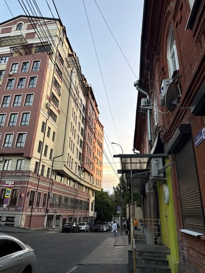 This is a low-angle, vertical shot of a city street in Sulak, Russia. The scene depicts a mix of old and new architecture, with several multi-story buildings lining the street. The sky is clear and appears to be either early morning or late afternoon, judging by the soft lighting.
In the foreground, a silver car is parked on the right side of the street, its rear end visible. The street itself is paved, with a sidewalk on the right. Along the sidewalk, there are several parked cars, including a black SUV. A few people are visible walking or standing near the buildings, dressed in casual clothing. One person in a white shirt and grey pants is walking away from the camera.
The buildings are varied in their design. On the left, a taller, modern building features a light-colored facade with many windows and visible air conditioning units. Further down the street, there are older brick buildings with decorative elements. Some of these buildings have storefronts at street level. Signs are visible on some of these storefronts, including one that reads "tōga" and another with a red circle and a blue cross inside a yellow diamond, indicating a traffic sign. Another sign appears to say "Slim Club."
The upper parts of some buildings show exposed brickwork and architectural details. Electrical wires are strung across the frame, adding a sense of urban density. The overall impression is a typical urban street scene in Russia, showcasing a blend of historical and contemporary architecture under a clear sky.