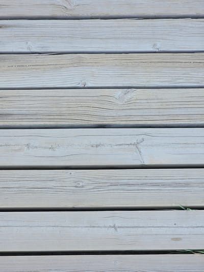 The image shows a close-up, top-down view of a weathered wooden surface composed of horizontal planks. The wood is a light grey color, with visible grain patterns and occasional knots. There are slight gaps between some of the planks, through which glimpses of a darker surface, possibly earth or gravel, are visible. A few blades of dry grass are present along the right edge of the frame, peeking through the gaps. The scene appears to be an outdoor environment, likely a boardwalk or deck. The image does not contain any people or specific identifiable landmarks that would indicate its precise location in Deveses, Spain.