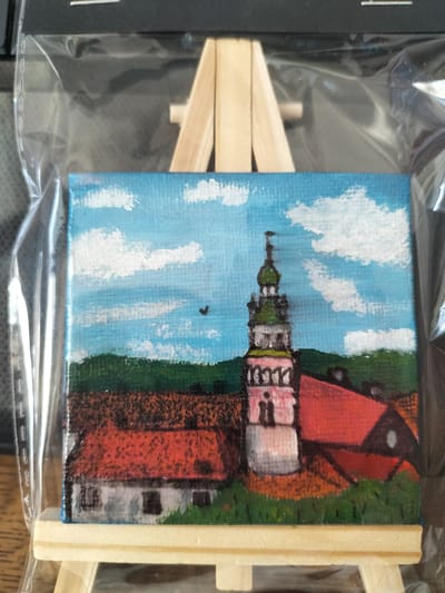 This is a small, square acrylic painting of a European town scene, likely Český Krumlov, Czechia, given the distinct tower. The artwork is enclosed in clear plastic packaging and displayed on a miniature wooden easel.

The painting depicts a bright blue sky filled with fluffy white clouds, suggesting a fair weather day. In the mid-ground, a lush green hillside forms a backdrop. Prominently featured in the right half of the painting is a tall, multi-tiered tower with a green, onion-shaped roof and a spire. The tower is painted in shades of pink, white, and gray, with arched window-like details. To the left of the tower and in the foreground, there are several buildings with red tiled roofs. One building on the far left appears to be a white house with dark windows. A smaller, darker building with a red roof is visible to the right of the tower. The ground in front of the buildings is depicted with patches of green grass and reddish-brown earth. A small, dark bird is flying in the sky above the green hillside.

The overall impression is that of a picturesque, possibly historic European town bathed in daylight. There is no visible text in the image.
