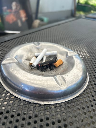 An ashtray sits on a black metal perforated surface. The ashtray contains several cigarette butts, including white-filtered cigarettes, a brown cigarette butt, and a black cigarette butt labeled "SOBRANIE." The ashtray is silver-colored and round. The location is Malinovo, Bulgaria.