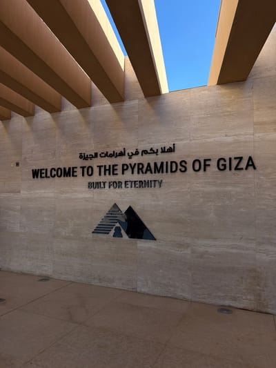 The image shows the entrance to the Pyramids of Giza, located in Kafrat Nassar, Egypt. A modern architectural structure with horizontal wooden beams is visible overhead, casting shadows on a light-colored, stone-tiled wall. The wall features prominent black lettering that reads "WELCOME TO THE PYRAMIDS OF GIZA" in English, and Arabic text above it, "أهلاً بكم في أهرامات الجيزة" which translates to "Welcome to the Pyramids of Giza". Below the English text, smaller dark letters spell out "BUILT FOR ETERNITY". A stylized, dark, triangular logo resembling pyramids is mounted on the wall below the text. The ground in front of the wall is paved with large, square tiles. The sky is a clear blue. No people are visible in the scene.