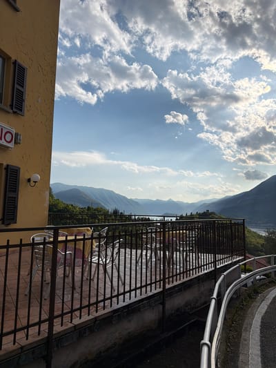 The image depicts a view from a balcony in Gittana, Italy. The balcony has a dark metal railing and overlooks a mountainous landscape. On the balcony, there are several metal chairs and tables with tablecloths. A yellow building with windows and shutters is visible in the lower left corner. Above the mountains, the sky is partially covered with white, fluffy clouds. A road with a railing is visible at the top right.