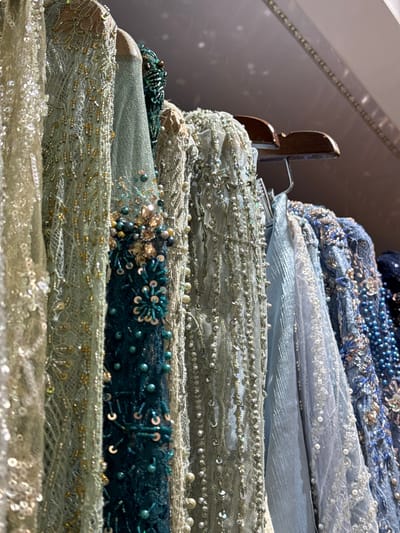 A close-up, low-angle shot showcases a row of intricately beaded and sequined garments hanging on wooden hangers. The fabrics, in shades of pale gold, mint green, teal, and various blues, are layered and textured, with a focus on the shimmering embellishments. The garments appear to be formal wear, possibly dresses or gowns, suggesting a boutique or a designer's studio. The background is blurred, but hints at a dimly lit indoor space, possibly a retail environment in the city of Tanjungmeru, Indonesia. The lighting highlights the sparkle of the sequins and beads, creating a luxurious and detailed visual.