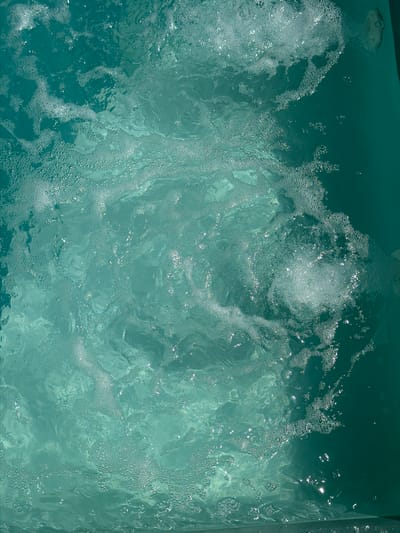The image shows the surface of a hot tub, filled with bubbling water. The water is a clear, light turquoise color with varying depths. Bubbles and foam are visible throughout the frame, suggesting the jets are active. The texture of the water's surface is rippled and disturbed. The location is not discernible from the image. No people or specific objects other than the water and tub are visible.