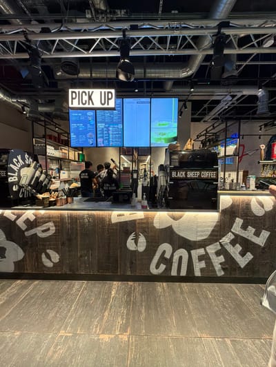 The image shows the interior of a "Black Sheep Coffee" shop. A brightly lit "PICK UP" sign hangs above a digital menu board displaying various drink options and prices. Behind the counter, three employees, two male and one female, are visible, appearing to be engaged in serving customers. The counter itself is made of dark wood with white lettering that reads "COFFEE" and a logo that looks like a sheep's head. On the counter, there are stacks of paper cups, a coffee machine, and various other items related to a coffee shop's operation. In the background, shelves are stocked with products. The floor is tiled. The image appears to be taken from the perspective of a customer standing in front of the counter. The presence of the "Black Sheep Coffee" branding and the general aesthetic suggest this is a commercial establishment. While the image depicts a coffee shop, there is no information within the image itself to specify its exact location within the city of Luton, United Kingdom.