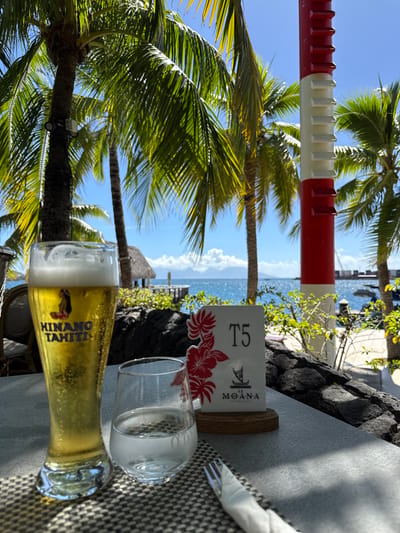 The image presents an outdoor dining table setting overlooking a tropical coastal environment. In the foreground, a tall glass of "HINANO TAHITI" beer with a foamy head is positioned on the left side of a dark patterned placemat. To its right, a shorter, stemless glass contains clear water. A white napkin partially obscures a silver fork to the right of the water glass. A white table sign, adorned with red floral patterns, displays "T5" at the top and "LE MOANA RESTAURANT" with a canoe logo at the bottom, standing on a wooden base. The table surface is light grey.

The midground features dark, textured rock landscaping separating the dining area from the water. Numerous lush green palm trees with visible trunks and fronds dominate the upper and central portions of the frame, indicating a sunny, outdoor setting. Beyond the rock wall, a thatched-roof bungalow is visible over blue water, connected by a wooden pier. The ocean extends to the horizon, with distant landmasses and mountains visible under a clear blue sky with scattered white clouds. On the right side, a tall, vertical red and white striped pole, featuring horizontal slats or vents, stands near the water's edge. Hints of a dock or pier with moored boats are discernible further right.

This scene is located at Le Moana Restaurant, part of the Hilton Hotel Tahiti in Papeete, France.
