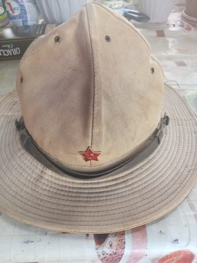 The image shows a close-up, top-down view of a tan-colored hat with a wide brim, possibly a boonie hat or a bush hat. The hat is placed on a surface that appears to be a tablecloth with a patterned design. A prominent feature on the hat is a red, five-pointed star emblem with a golden hammer and sickle inside, which is a symbol often associated with the Soviet Union. The hat also has a dark strap around its crown, fastened with metal clasps. Two dark grommets are visible on the side of the crown, likely for ventilation. The fabric of the hat appears to be canvas or a similar durable material, and it shows some signs of wear, with minor dirt marks.

The scene is indoors, as suggested by the lighting and the presence of items that appear to be from a household or a shop. To the left of the hat, parts of some cylindrical objects, possibly cans or containers, are visible. One label shows the word "ORACLE" and "Choice" in dark text on a brown background. Another label shows the word "Rondal" in yellow text on a white background. The overall lighting suggests it might be daytime, but the exact time and weather conditions are not discernible. The setting is not specifically identifiable as Мариновка, Kazakhstan, but the context provided suggests this as the location. There are no people in the image, and the focus is entirely on the hat and its surroundings. The image conveys a sense of historical or political interest due to the presence of the Soviet star emblem.