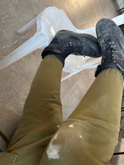 An individual's legs are visible, clad in olive drab pants and black combat boots, resting on a white plastic chair. The chair is positioned on a floor tiled with light brown and gray marbled tiles. The scene appears to be indoors, possibly in a public area or facility within the Eshkol Regional Council, Israel, given the characteristic architecture and materials often found in such locations. The boots and pants show signs of wear and dirt, suggesting outdoor activity or work.