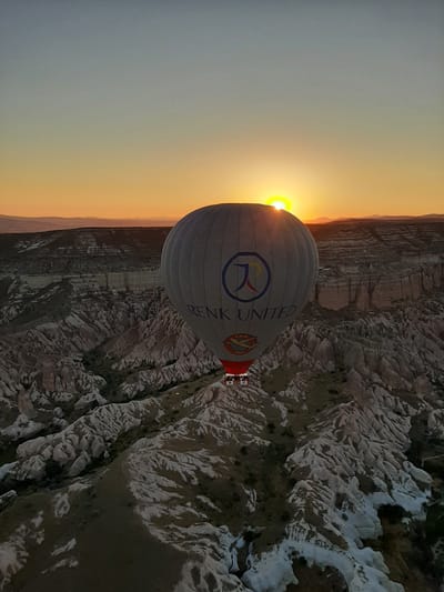 A hot air balloon is captured mid-flight over the unique geological formations of Cappadocia, near Avanos, Turkey, during sunrise. The sun is peeking over the horizon, casting a warm, orange glow across the sky and illuminating the landscape. The balloon, predominantly white with blue and red lettering that reads "RENK UNITED" and a circular logo featuring an abstract design and a small airplane, is the central subject. A basket carrying several passengers is suspended beneath it. The terrain below consists of a dramatic valley filled with weathered rock pillars and formations, common to the region. The sky transitions from a pale blue at the top to a vibrant orange and yellow gradient near the horizon.