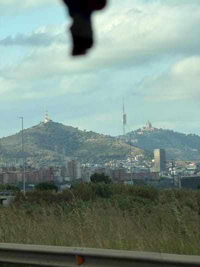 The image captures a panoramic view of the city of El Prat de Llobregat, Spain, from a slightly elevated vantage point. In the foreground, a blurred, dark object hangs from the top of the frame, obscuring a portion of the sky. Below this, a metal guardrail runs horizontally across the bottom of the image, suggesting the photograph was taken from a vehicle or a bridge.

The middle ground is dominated by a dense expanse of tall, dry grasses and green vegetation, indicative of an uncultivated area or roadside verge. Beyond this natural landscape, the urban sprawl of El Prat de Llobregat unfolds. Numerous residential buildings, varying in height and architectural style, form a complex cityscape. Some buildings are constructed with reddish-brown bricks, while others feature lighter facades. Amidst the urban structures, several taller, modern buildings stand out, including a prominent, tall, rectangular skyscraper.

In the background, rolling hills rise above the city. The most distinctive features on these hills are two large towers and a cluster of buildings resembling a complex on a hilltop. One of the towers, tall and slender with a lattice structure, is clearly visible. The other tower, located on a separate, closer hill, is topped with a red and white structure. The hilltop settlement appears to have a prominent architectural design, possibly a church or historical site, with multiple spires. The sky above is partly cloudy, with soft, diffused light illuminating the scene.