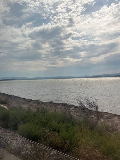 This media file depicts a scenic view of a body of water, possibly a lake or the sea, with land and a cloudy sky. The water is bright and reflects the light, suggesting it is daytime. A shoreline with sparse vegetation and some dry, bare branches is visible on the right. In the distance, a hazy outline of mountains or hills can be seen along the horizon. The sky is filled with a mix of light and dark clouds, with streaks of sunlight breaking through. The overall impression is one of natural beauty and tranquility, possibly during daytime under a partly cloudy sky. There is no visible text in the image. The location context is Narbonne, France.