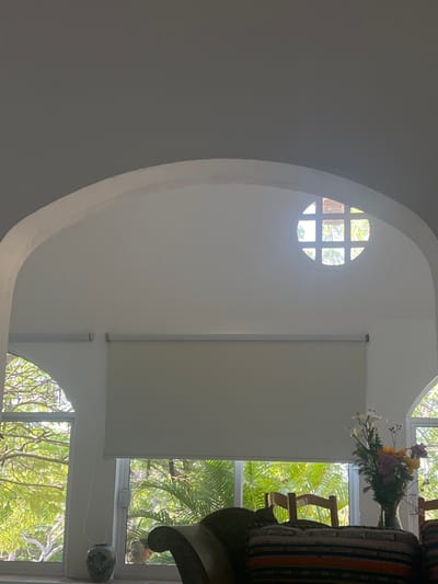 An interior view showcasing architectural details, including a curved wall with a small, round, multi-paned window. A large window with a white blind is visible, revealing lush greenery. A vase of flowers sits near the window, next to a partially seen armchair with colorful pillows. Another arched window is located in the bottom right corner. The ceiling and walls are primarily white.