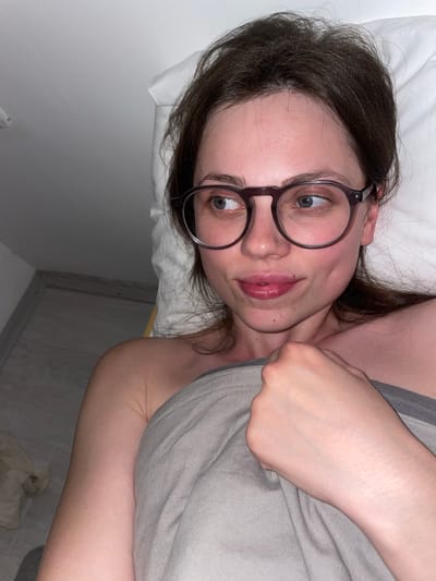 A woman with dark hair and round glasses is lying in bed. She is partially covered by a gray blanket. She is looking up at the camera with a slight smile. The background is a white pillow and wall. The location is not discernible from the provided image.