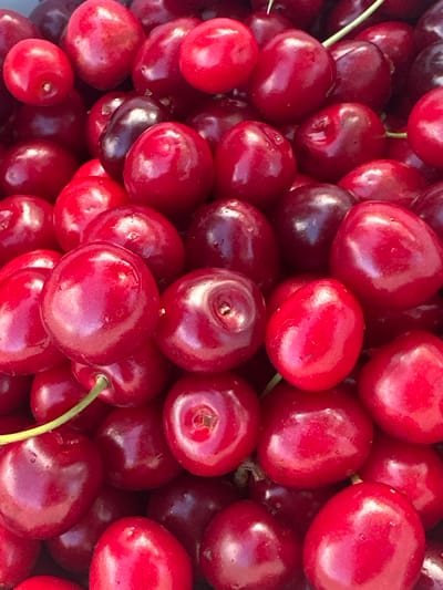 A close-up, overhead shot showcases a large pile of ripe cherries. The cherries are a vibrant red, with some appearing slightly darker, almost purplish, due to shadows and their inherent color variations. They are clustered tightly together, with their smooth, glossy skins reflecting light. Several stems are visible, some green and fresh, others darker. The composition fills the frame, creating an abstract pattern of round shapes and deep red hues. This image was taken in Höslwang, Germany.