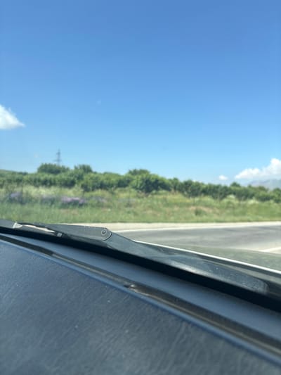 The image provides a view from inside a vehicle, looking out through the windshield. The foreground is dominated by the dark, textured surface of the car's dashboard, with a windshield wiper blade visible extending across the lower portion of the frame. Beyond the windshield, a landscape unfolds under a clear, bright blue sky. A row of lush green trees and vegetation forms a dense band across the middle ground, with patches of purple wildflowers interspersed within the greenery. A power line tower is faintly visible behind the trees on the left side of the frame. A light-colored road curves to the right in the distance. The image appears to be taken on a sunny day. There is no clear indication of the city of Zangakatun, Armenia, within the image itself.