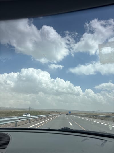 A passenger perspective of driving on a highway outside Talin, Armenia, featuring a clear blue sky with large, fluffy white clouds. The highway appears to be a multi-lane road with a yellow divider line and guardrails. A white car is visible on the side of the road. The landscape beyond the highway consists of dry, grassy terrain.