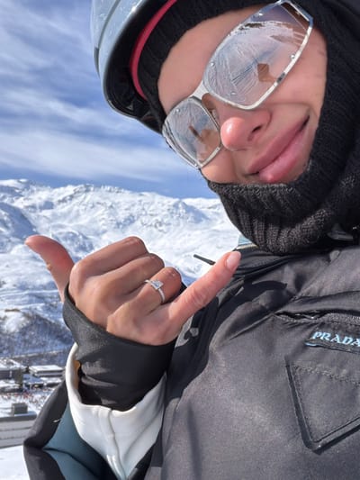 A close-up, low-angle selfie depicts an individual positioned on a snow-covered mountain, likely within a ski resort in the commune of Les Belleville, France. The person wears a grey ski helmet with a red trim, a black balaclava covering the lower face, and silver-framed rectangular sunglasses displaying condensation and reflections of a handheld device. The individual's right hand is raised, forming a "shaka" gesture, prominently featuring a silver-colored ring with a princess-cut diamond solitaire and pave-set diamonds on the band, worn on the ring finger. The individual is dressed in a black insulated jacket with a light blue "PRADA" logo embroidered on the left chest, over an off-white undergarment visible at the wrist. The background reveals vast, snow-covered mountain ranges under a clear blue sky, with ski resort buildings and activity observed in the valley below.