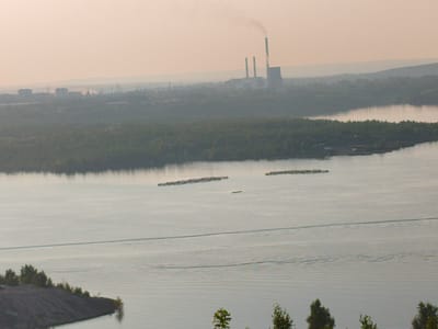 An aerial view shows the city of Veselyy, Russia, bathed in the soft light of either sunrise or sunset. The scene is dominated by a wide body of water, likely a river or lake, reflecting the pale, hazy sky.
In the distance, a sprawling industrial complex is visible, characterized by tall smokestacks emitting plumes of white smoke into the air, a clear indication of active industrial processes. Structures that appear to be factories or power plants dot the landscape, blending into the hazy atmosphere.
Closer to the foreground, the banks of the waterway are lined with dense green trees, forming a lush natural border. The water itself is marked by the presence of several clusters of buoys or floating structures, arranged in long lines, suggesting a possible aquaculture or boating area. A lone, small boat is visible on the water, leaving a faint wake.
On the left side of the image, a prominent embankment or hill composed of dark, granular material rises from the water's edge, sparsely vegetated with trees. The overall impression is one of a landscape where industrial activity and natural elements coexist, softened by the diffused atmospheric light.