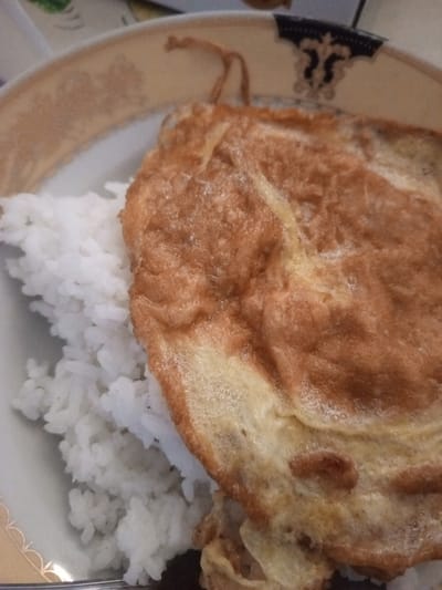 A close-up, slightly overhead shot shows a plate of white rice topped with a golden-brown omelet. The rice is fluffy and piled high on the left side of the plate, while the omelet, which appears to be cooked evenly with some slight browning and a bubbly texture, covers most of the right side. The plate itself is white with a decorative gold trim and a darker blue and black pattern around the rim. The image is taken in Mvera Mission, Malawi, where this simple yet appetizing meal is being served.