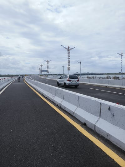 An outdoor, eye-level, wide shot captures a view of the Bali Mandara Toll Road in South Denpasar, Indonesia, under an overcast sky. The dark asphalt road dominates the foreground, with a bright yellow line marking the lane. Concrete barrier walls line the right side of the road, and on the left, white and red barriers indicate the edge of the road. Several motorcycles are traveling in the distance on the left lane, and a silver minivan is moving forward on the right lane. Tall, ornate streetlights with decorative elements and lights stand along the right side of the toll road, interspersed with modern signposts. The background shows a body of water and a distant bridge. The overall atmosphere is one of an active, modern infrastructure project.