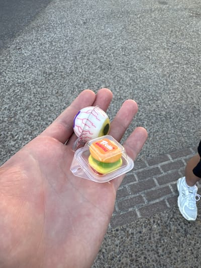 In this close-up, outdoor shot, a person's open hand holds two Halloween-themed candies. The hand is positioned over a textured, pebbly surface, likely a sidewalk or pathway, with a brick pattern visible in the background. The lighting suggests it's daytime, with a shadow cast across the brickwork.
The candies are a novelty eyeball candy with pink veins and a purple iris, and a small clear plastic container holding two gummy candies. The container has the "Trolli Originals" logo printed on its lid in orange and white lettering. One of the gummies appears to be lime green, and the other is a bright orange.
In the background, parts of two people are visible. One person is wearing a light-colored athletic shoe with white laces, and the other person is wearing dark pants and the same type of shoe. The overall scene conveys a casual, possibly festive atmosphere, suggesting the candies might be for an event like Halloween. The setting appears to be outdoors in an urban or park environment, given the paved surfaces.