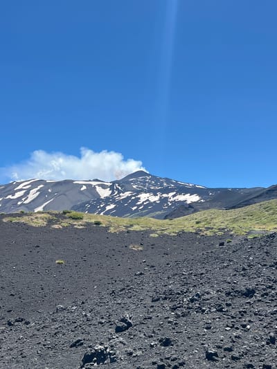 The image shows a volcanic landscape near Linguaglossa, Italy. In the foreground, there is a desolate area covered in black volcanic rock and gravel. Further back, a grassy slope with sparse vegetation leads up to a snow-capped mountain. The mountain appears to be a volcano, with a plume of smoke rising from its summit. The sky is clear and blue. The image is a photograph taken from a low angle, looking up at the volcano.
