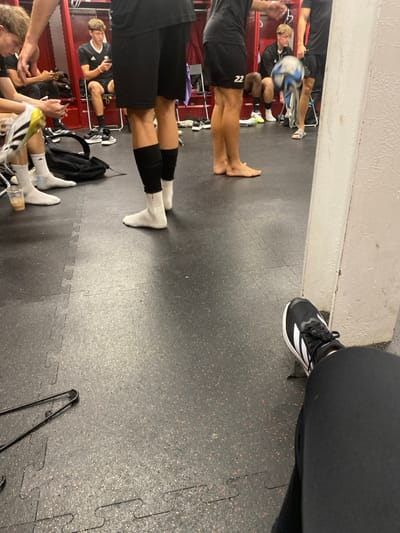 The image depicts an indoor locker room scene in Lawrence Township, United States. The room features a red locker bank along the rear wall and a black interlocking rubber mat floor. Several young males, attired in athletic wear including black shorts, jerseys, and socks, are present. Some are seated on benches, others stand. In the mid-ground, one individual is partially visible, juggling a black and white soccer ball. Another person stands barefoot, revealing a small tattoo on the right ankle. Other individuals are observed holding or looking at mobile phones. A black duffel bag, a plastic cup containing a beverage, and assorted athletic footwear, including cleats and sandals, are visible on the floor. In the immediate foreground, the legs and feet of the person capturing the image are visible, wearing black athletic shoes with white stripes and black athletic pants. A metal hanger or rack component rests on the floor in the lower left.