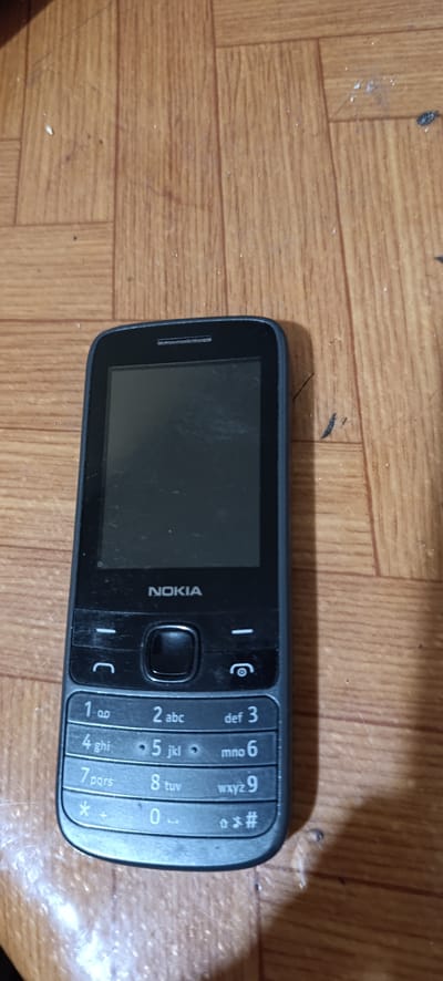 The image shows a black Nokia feature phone lying on a wooden surface. The phone is positioned vertically, with the screen facing upwards. The screen is dark, indicating it is either off or displaying a black background. The Nokia logo is visible just below the screen, in white text. Below the logo is a set of navigation buttons, including a central circular button. The numeric keypad is also visible, with numbers 1 through 9, 0, *, and # clearly marked. The keys also show the corresponding letters for dialing. The wooden surface appears to be a table or floor, with a visible grain pattern and some light reflections. There are also some small white specks and dust particles on the surface, suggesting a lived-in environment. The lighting appears to be from an overhead source, casting soft shadows. No people are visible in the image. The context of Svetlen, Bulgaria is not directly evident from the visual cues in the image itself, but implies a domestic or everyday setting. The overall impression is a still life of a simple, functional mobile device.