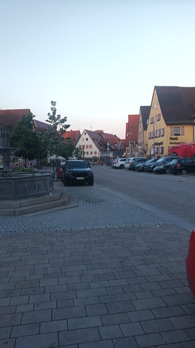 This image depicts a street scene in Altdorf bei Nürnberg, Germany, during the late afternoon or early evening, as indicated by the soft, warm light and long shadows. The sky is a pale blue, transitioning to a lighter hue near the horizon.

The foreground features a cobblestone plaza with a large, ornate fountain to the left. Beyond the plaza, a row of parked cars lines the street. The cars are a mix of makes and models, including a black SUV prominently parked in the center, a red car visible on the far right, and several white and gray vehicles.

The buildings lining the street are characteristic of European architecture, with pitched red-tiled roofs and facades featuring a mix of colors, including yellow and white. Some buildings have visible windows with shutters. One building displays signage for "Hotel Fichtenberger Hof". There are also red umbrellas visible outside one establishment, suggesting an outdoor seating area.

The scene appears quiet, with no visible people actively engaged in activities, though the presence of parked cars and outdoor seating implies a community or business district. The overall mood is serene and peaceful, characteristic of a European town square in the twilight hours.
