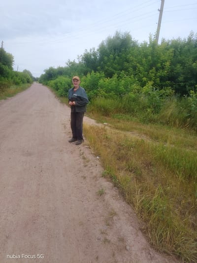 This is a photograph taken outdoors, likely in a rural area, possibly in Sestrynivka, Ukraine, given the context. The scene depicts a dirt road stretching into the distance, flanked by lush green trees and vegetation. The sky is overcast, suggesting a cloudy or possibly rainy day, and the time of day appears to be daytime.
The central subject of the image is an elderly man standing on the right side of the dirt road, facing the camera. He is wearing a light brown cap, a dark green jacket over a red shirt, and dark trousers. His posture is relaxed, with his hands clasped in front of him. He has a slight smile on his face, conveying a sense of mild contentment or perhaps a friendly acknowledgment of being photographed.
To the left of the man and stretching out to the horizon is the unpaved road, appearing to be a country lane or a path through a wooded area. The road surface is light brown and dusty, with scattered small stones. On the right side of the road, a thick growth of grass and various plants can be seen, some of which are dry and yellow, indicating possibly late summer or early autumn. Beyond the vegetation, more trees form a dense backdrop.
In the background, a utility pole with power lines is visible on the right side, suggesting the presence of infrastructure even in this seemingly remote location. The overall mood of the photograph is peaceful and serene, capturing a simple moment of a person in nature. The lower left corner of the image displays the text "nubia Focus 5G".