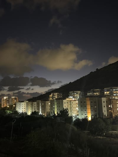 The image depicts a nighttime view of the city of Tirat Karmel, Israel, with a focus on residential buildings situated on a hillside. The sky is dark, with scattered clouds illuminated by artificial light, creating a dramatic contrast. Several multi-story apartment buildings are visible, their windows lit, indicating occupancy. A prominent dark, tree-covered hillside dominates the right side of the frame, casting a shadow over some of the buildings. In the foreground, there is a dark, vegetated slope with some low-lying vegetation and a solar-powered lamp post. The overall impression is one of a residential area at night, with the natural landscape of the hillside providing a backdrop to the urban development.