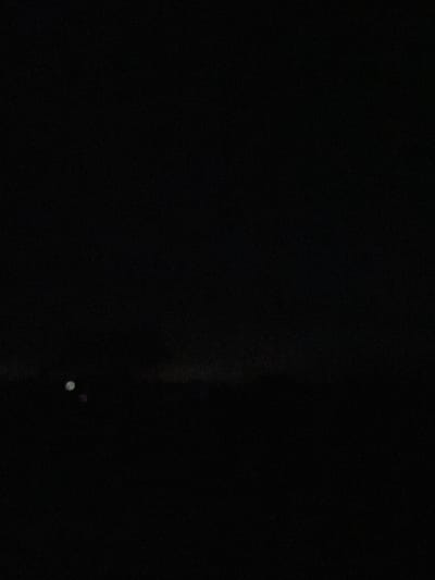 The image is an underexposed nighttime photograph, predominantly black, suggesting a very dark environment. In the lower left quadrant, there are two faint, out-of-focus circular lights. One is a pale white, and the other is a slightly dimmer, more diffused light. The background appears to be a dark, indistinct horizon line with very subtle hints of lighter shades, possibly indicating distant, low-lying terrain or structures against a dark sky. The overall impression is one of minimal visibility. The location of Yermekeyevo, Russia, is not visually represented in this image.