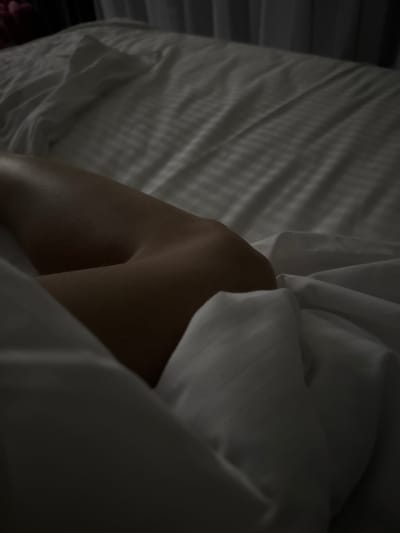 The image shows a close-up, dimly lit view of a person's leg, specifically the thigh and knee area, resting on white sheets in a bed. The sheets have a subtle striped texture. The person's skin appears smooth and is illuminated by soft, indirect lighting, casting gentle shadows that highlight the contours of the leg. The background is out of focus and dark, with faint vertical lines suggesting curtains or blinds. The overall impression is one of intimacy and quiet repose. The location information "Doma 1096 Km, Russia" is a place name and not descriptive of the visual content of the image.