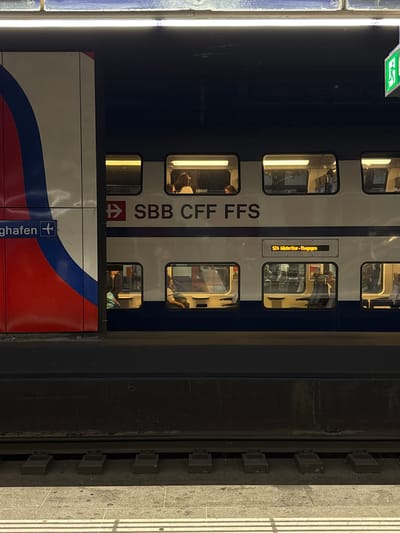 A double-decker SBB CFF FFS train is stationary at a platform within an indoor railway station, likely Zurich Airport railway station in Kloten, Switzerland, indicated by a partial "ghafen" sign with an airplane icon on a left-side wall panel. The train is predominantly white with a dark blue lower section, featuring multiple rectangular windows on both decks. An amber LED display on the train's side indicates "S24 Winterthur - Thayngen." Through the illuminated windows, several passengers are visible seated inside the train on both levels. The platform consists of grey concrete with white tactile paving stripes along its edge, bordering two parallel railway tracks. Overhead, fluorescent lights illuminate the station, and a green exit sign is suspended from the ceiling on the right side of the frame.