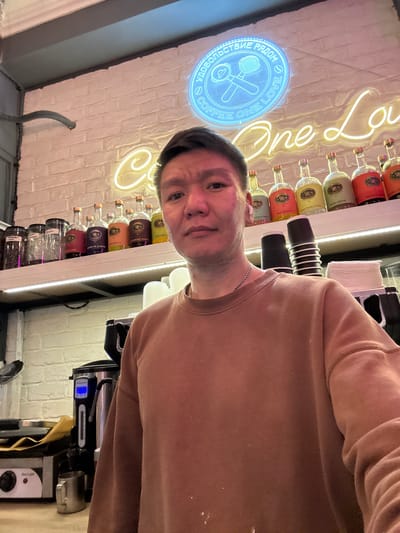 A man is taking a selfie in a brightly lit cafe. He is wearing a brown sweatshirt and a silver chain. Behind him, a white brick wall is adorned with neon signs. A circular blue neon sign with text and a coffee-making apparatus is above and to the left of a larger, yellow neon sign that reads "One Love". Below the signs, bottles of various colored liquids are displayed on a shelf. To the right of the man, stacks of black and white disposable cups are visible. Further to the right, a coffee machine is partially visible. On the counter in the foreground to the left, there is a hot plate with a knob labeled "Hot Light" and a small metal pitcher. The cafe appears to be located in Veshki, Russia.