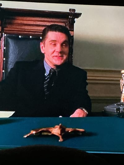 A man in a suit and tie is seated behind a desk in what appears to be an office. He is looking towards the camera with a slight smile. In front of him on the desk is a small, reddish-brown object that resembles a dried leaf or a piece of bark. The background shows a wooden paneled wall and a large, ornate chair. The overall lighting is somewhat dim, suggesting an indoor setting. The specific location in Hallandale Beach, United States of America, cannot be determined from the visual information provided in the image.