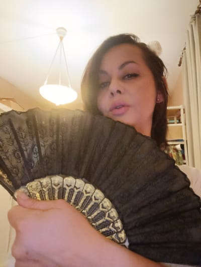 A woman is holding a black lace fan in front of her face in an indoor setting. She is looking at the camera with a neutral expression. The background is blurred but appears to be a room with a ceiling light fixture and curtains. There is no indication of the city of Serravalle, Switzerland in the image.