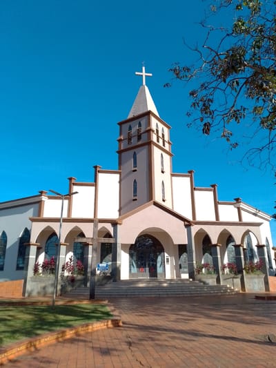 A photograph, taken from a low angle, captures the front facade of a church under a clear, bright blue sky. The church is a light-colored building with a prominent bell tower topped with a cross. The tower has several arched windows and decorative trim. The main body of the church features a series of arched entrances and windows, with a pinkish-tan facade. A large wooden cross stands in front of the entrance. The foreground is paved with reddish-brown bricks, and a strip of green grass with a brick border runs along the left side of the paved area. Reddish-purple plants are visible in planters along the front of the church. A street lamp stands to the left of the main entrance. The image appears to be taken during daylight, casting shadows on the ground. There are no people or animals visible in the frame. This church is identified as Igreja Evangélica Assembléia de Deus in São Gabriel Do Oeste, Brazil.