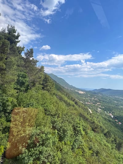 The image shows a landscape view, likely taken from an elevated position in or near Čavori, Montenegro. The sky occupies a significant portion of the frame and features a bright blue color with scattered white clouds. Below the sky, a mountain range is partially visible, with a portion of the Adriatic Sea appearing in the distance. The slope of a densely forested mountainside is the primary subject. The foliage is predominantly green, indicating lush vegetation. Scattered buildings and residences are visible on the mountainside. There is a visual obstruction on the left side of the sky.