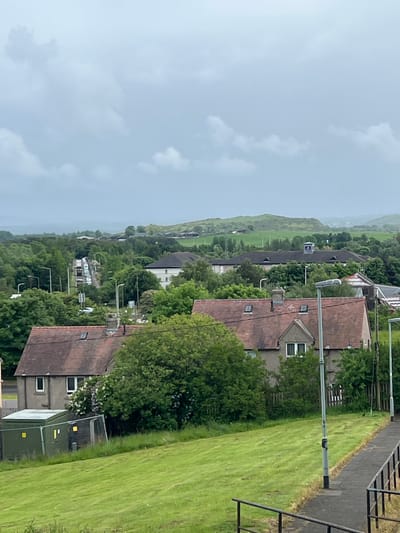 The image displays a panoramic view of a residential area transitioning into a rural landscape in Blackburn, United Kingdom, under an overcast sky. The upper portion of the frame shows a light blue-grey sky with soft, diffuse clouds, indicating cloudy daytime conditions.
In the mid-ground and foreground, a cluster of residential buildings is visible, characterized by their reddish-brown, pitched tiled roofs and light-colored facades. These homes are interspersed with lush green trees and well-maintained grassy areas, giving a verdant appearance to the neighborhood. A paved path or road runs along the top right edge of the frame, lined with modern streetlights and a dark metal railing. Further into the residential sprawl, other structures, including what appears to be a larger, darker-roofed building with a small white tower, are nestled amongst the trees. Some cars are partially visible parked on a street further back, obscured by foliage. A green utility box with indistinct yellow labels can be seen close to one of the houses.
Beyond the immediate housing, the landscape opens up to expansive, rolling green hills in the distance, covered in trees and fields, suggesting a proximity to natural countryside. The overall scene depicts a calm, residential setting integrated with its natural surroundings. No people or specific activities are discernible in the static landscape. There is no legible text visible within the image.